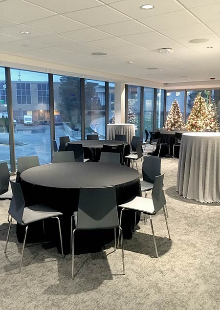 An image of the Spencer Theatre Donor Lounge, a large room with full-length windows the southern and western walls, with a white pop-tile celeing, and carpeted floors. The room is decordated for a Christmas party: three Christmas trees decorated with white lights are in the background, and in the foreground are party tables. 3 of the party tables have black tablecloths with 8 black chairs around each of them. 2 of the tables are high-tops with silver tablecloths. 