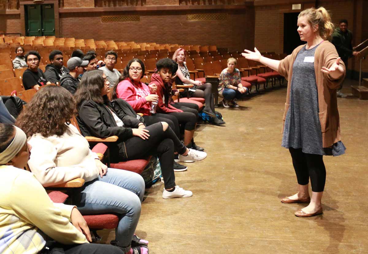 Nicole Marie Green, a white woman with blonde hair pulled back into a ponytail, stands with her arms open wide as she talks to a diverse group of highschool students. Nicole wears a dark grey dress over black leggings, brown ballet flats, and a long brown cardigan with pockets that is almost the same length as her dress. There is a white nametag on her dress. The students she talks to are seated.