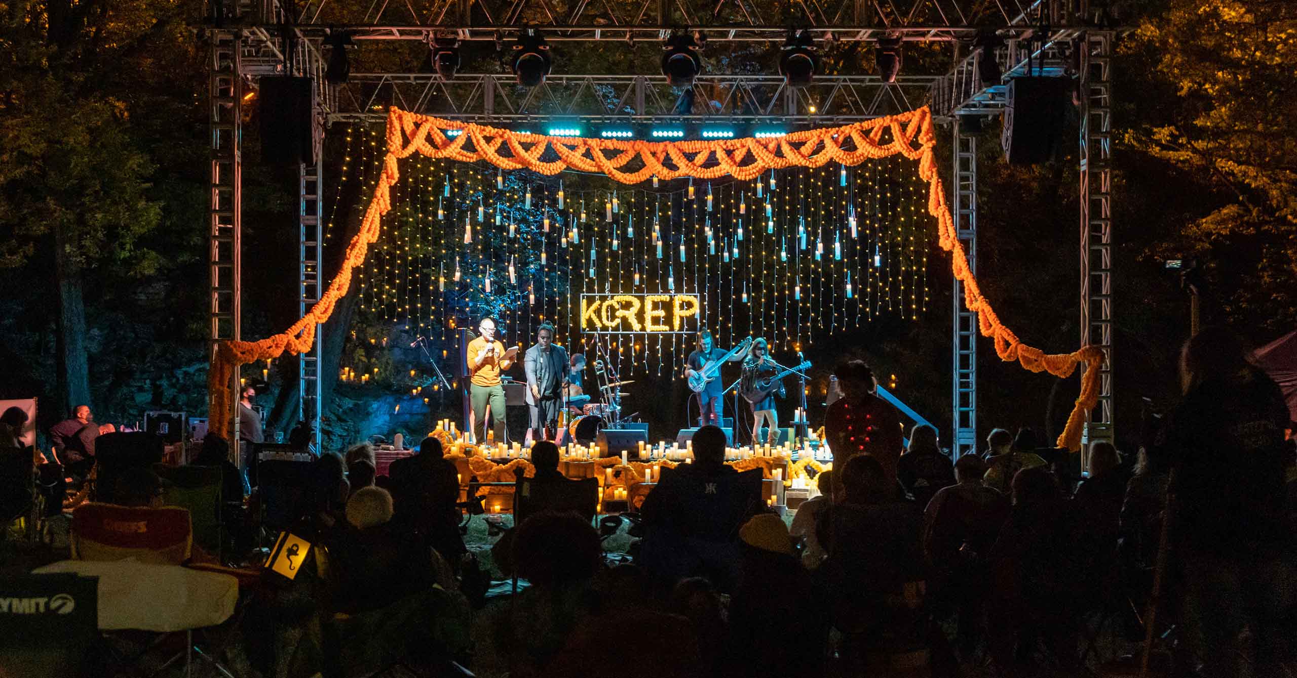 Stuart Carden, a white man with glasses in an orange shirt and green pants, introduces the musical act Deshica Rage to the Ghost Light stage - an outdoor stage decorated with candles and marigolds. It is nighttime and people seated in folding chairs make up the crowd.