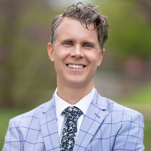 A photo of a white, middle aged man with sandy hair and wearing a blue checked suit. He smiles at the camera.