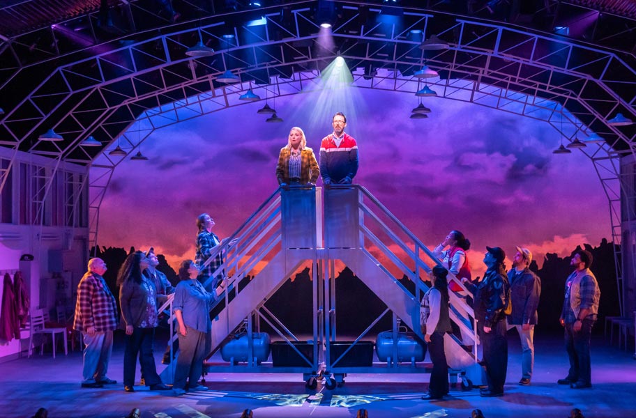 A theater set where a white woman in a plaid jacket and white man in a striped pullover stand on airplane stairs in an airplane hanger, surrounded by a crowd.