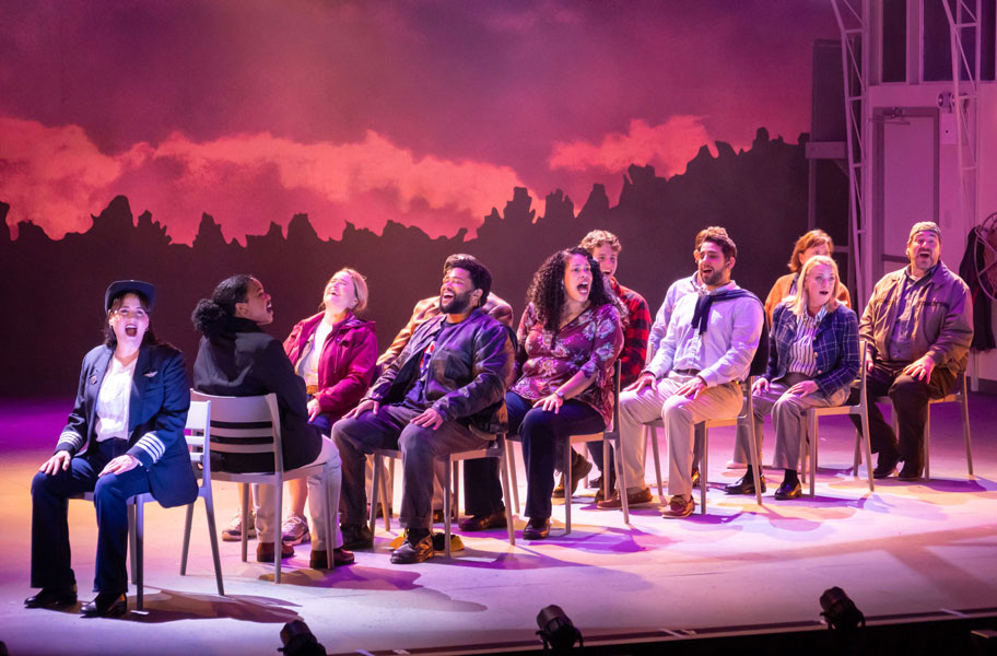 A theater set: onstage we see a group of people sitting on chairs arranged as though they are on an airplane with a person dressed as a pilot at the front of the group.